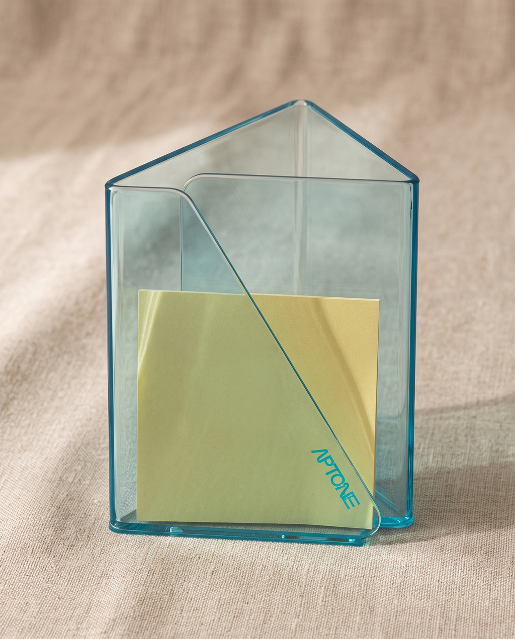 A transparent light blue house-shaped sticky note holder containing a pale yellow sticky note, set against a neutral beige fabric background.