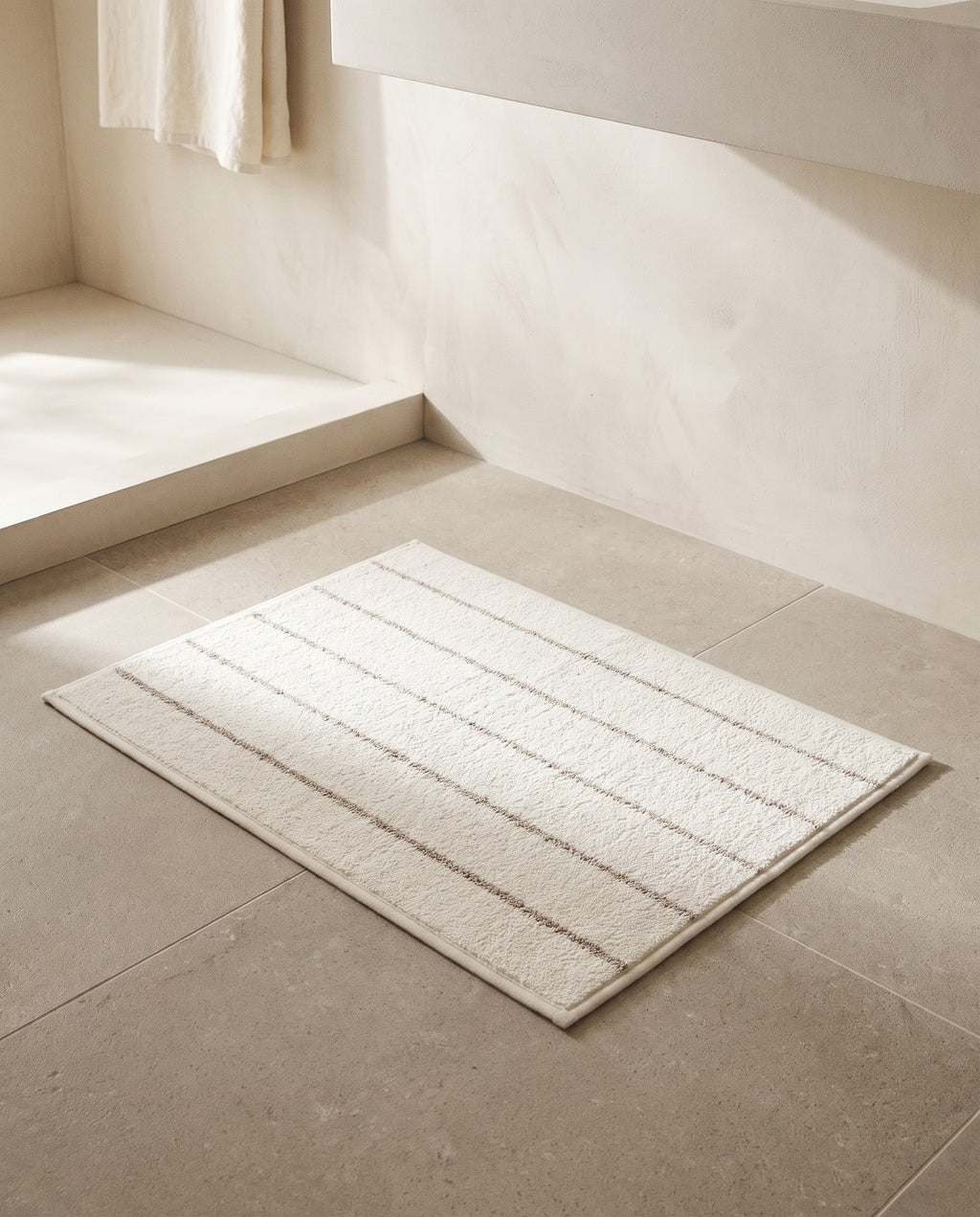 Beige stripe bath mat lying flat on a limestone bathroom floor in morning light