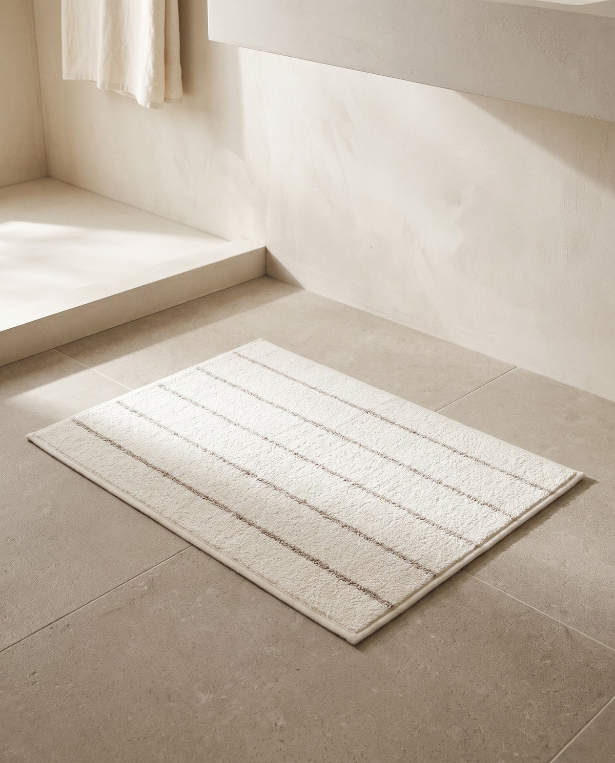 Beige stripe bath mat lying flat on a limestone bathroom floor in morning light