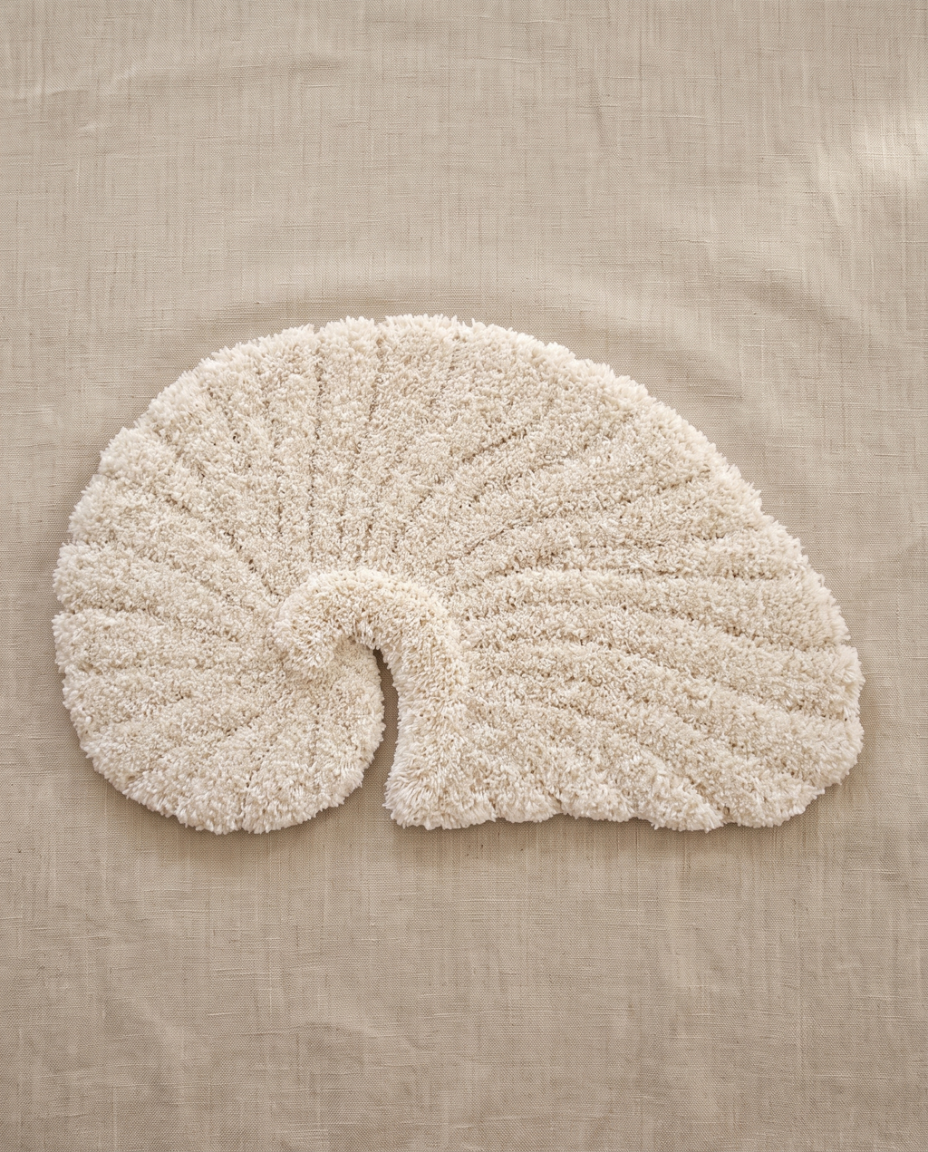 Poh object rug by Lug Lug in beige, a warm ivory nautilus shell tufted in shaggy pile, laid flat on natural linen