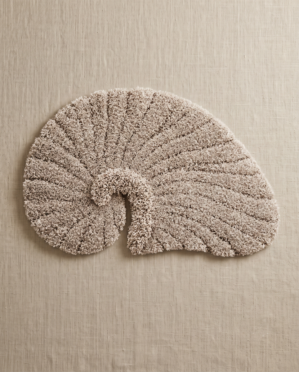 Poh object rug by Lug Lug in gray, a warm greige nautilus shell with the ribbed grooves reading sharply, laid flat on natural linen