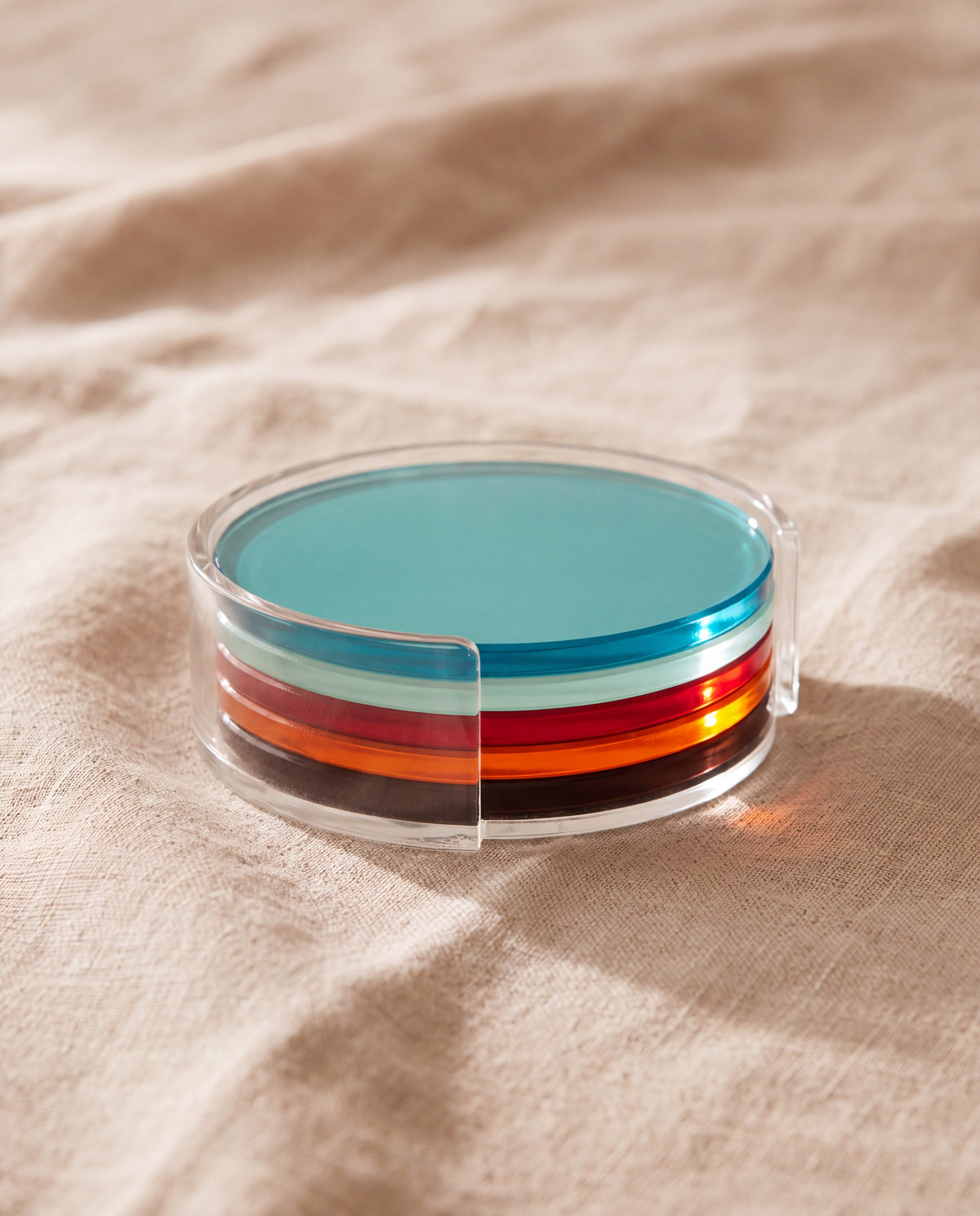Rainbow coaster by APTONE, five translucent acrylic discs stacked in their clear holder on beige linen