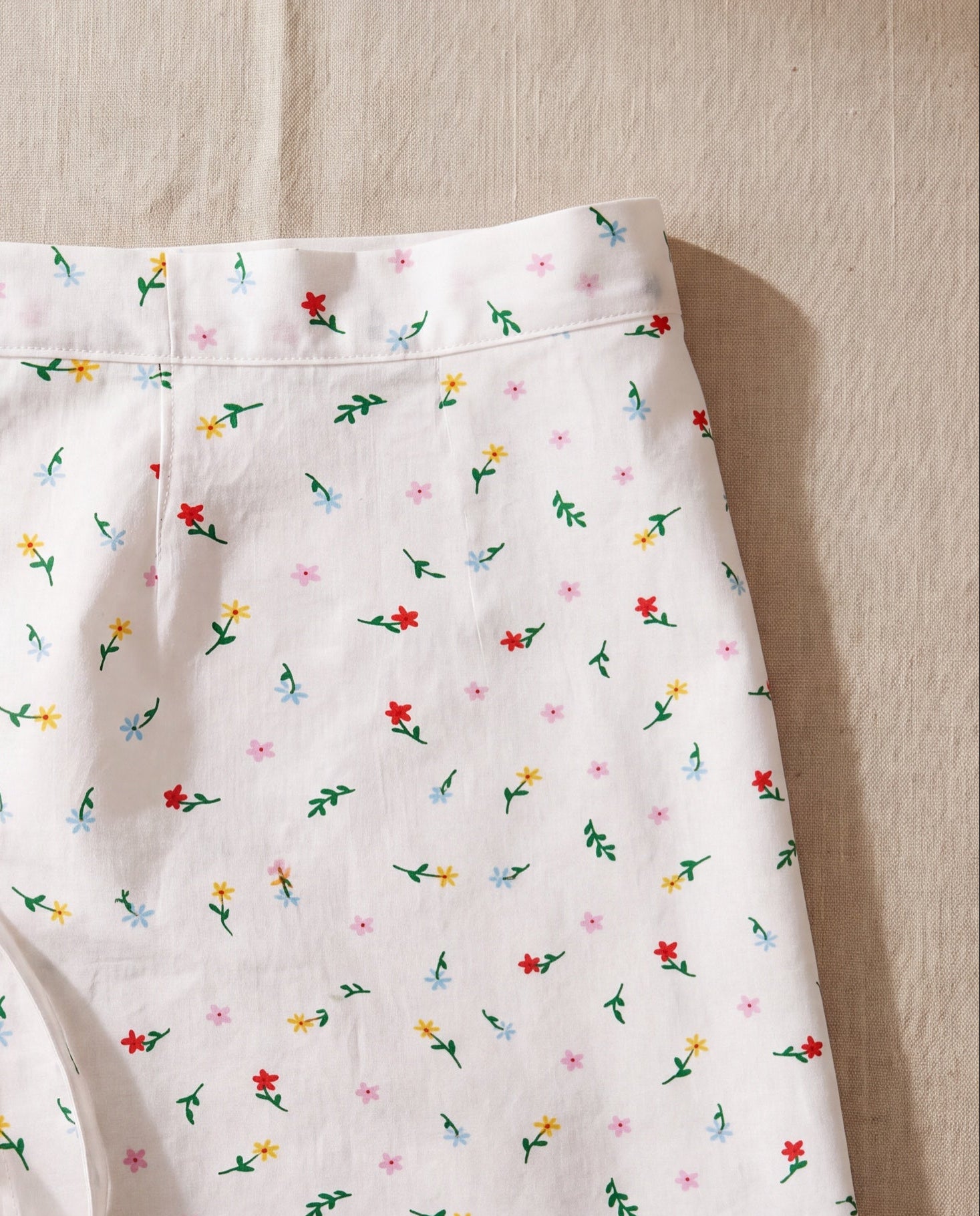Roa half apron by lemonbaseball livingclub, white cotton in a small scattered floral print, laid flat on beige linen
