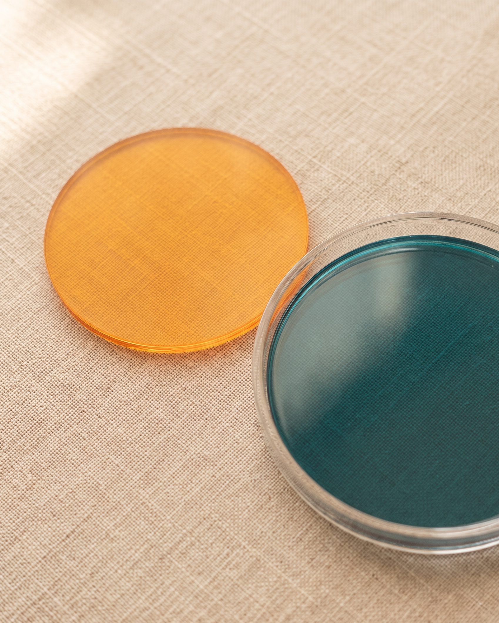 Rainbow coaster by APTONE, an amber disc on linen beside a turquoise disc resting in the clear acrylic holder